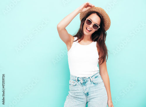 Young beautiful smiling female in trendy summer clothes. Sexy carefree woman posing near blue wall in studio. Positive brunette model having fun. Cheerful and happy. In hat