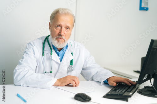 Portrait of serious mature adult male doctor in white coat with stethoscope working on desktop computer sitting at desk in medical office, looking at camera. Confident senior physician at workplace.