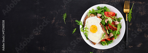 Breakfast - toast with fried eggs and tomatoes salad with greens and cod liver on dark background. Healthy breakfast. Top view, banner