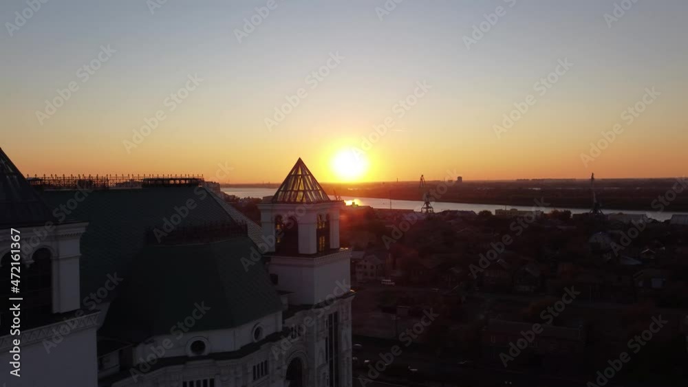Aerial view ascending over the city at golden hour, with the sun in the distance setting and creating a beautiful lens flare. An old white building with a glass tower.
