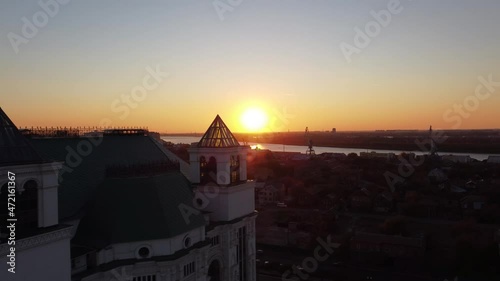 Aerial view ascending over the city at golden hour, with the sun in the distance setting and creating a beautiful lens flare. An old white building with a glass tower.