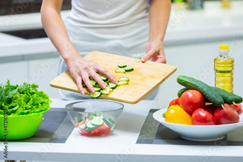 Close up of a girl hands slicing vegetables with a knife on a cutting board for a vegan vitamin vegetable salad and putting them in a glass bowl while cooking breakfast in the kitchen