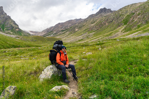 Tourist-mountaineer middle-aged man with a large backpack on his shoulders sits resting on a large rock against the background of green mountains