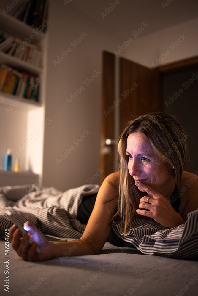 Obraz premium Pretty, middle-aged woman using her cell phone in bed at home before sleep
