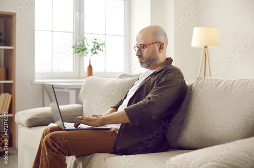 Serious bald bearded adult man in glasses sitting on comfortable sofa in living room at home, using modern laptop computer, watching educational video or working on online business project