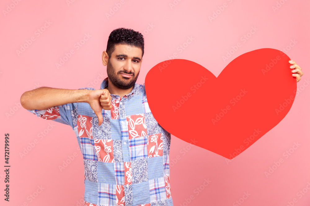 Upset young adult dark haired bearded man with sad facial expression holding big red heart in hands and showing thumb down, unrequited love. Indoor studio shot isolated on pink background. Upset young adult dark haired bearded man with sad facial expression holding big red heart in hands and showing thumb down, unrequited love. Indoor studio shot isolated on pink background.