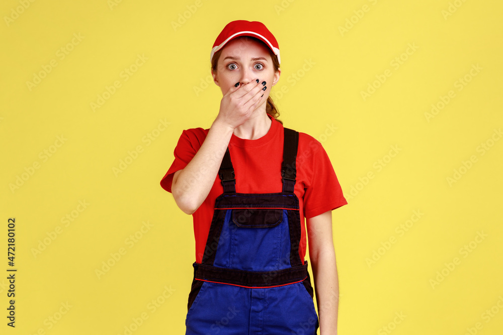 Portrait of shocked amazed worker woman standing and looking at camera with big eyes, covering mouth with palm, wearing work uniform and red cap. Indoor studio shot isolated on yellow background. Portrait of shocked amazed worker woman standing and looking at camera with big eyes, covering mouth with palm, wearing work uniform and red cap. Indoor studio shot isolated on yellow background.