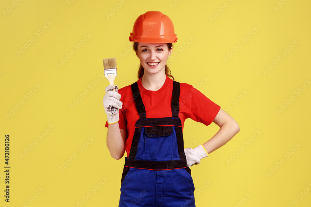Positive young adult builder woman standing with paintbrush in hands, wearing work uniform and protective helmet, keeps hand on waist. Indoor studio shot isolated on yellow background.