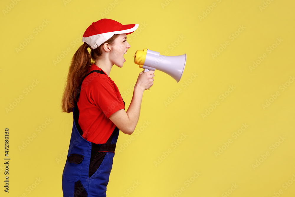 Side view portrait of angry worker woman screaming loud in megaphone, giving command her subordinate builders, wearing work uniform and red cap. Indoor studio shot isolated on yellow background. Side view portrait of angry worker woman screaming loud in megaphone, giving command her subordinate builders, wearing work uniform and red cap. Indoor studio shot isolated on yellow background.