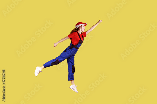 Side view portrait of woman worker flying to do her work like superhero, fast and high quality service, wearing work uniform and red cap. Indoor studio shot isolated on yellow background.