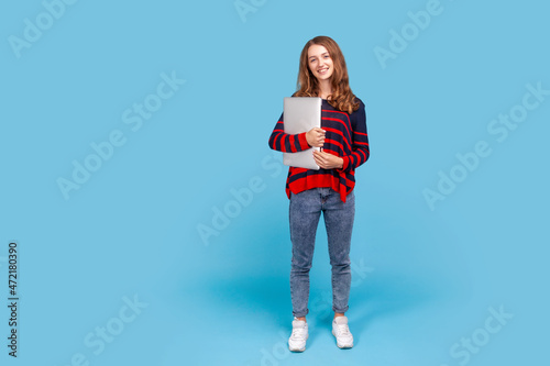Full length portrait of smiling woman in striped casual sweater, standing holding closed laptop or folder, looking at camera with positive expression. Indoor studio shot isolated on blue background.