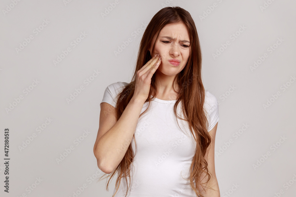 Unhealthy sick woman touching cheek with expression of terrible suffer from toothache, sensitive teeth, cavities, wearing white T-shirt. Indoor studio shot isolated on gray background. Unhealthy sick woman touching cheek with expression of terrible suffer from toothache, sensitive teeth, cavities, wearing white T-shirt. Indoor studio shot isolated on gray background.