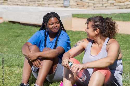 Content plus size women resting after training. Chubby women of different nationalities in sport clothes drinking bottled water, sitting on grass. Sport, body positive concept
