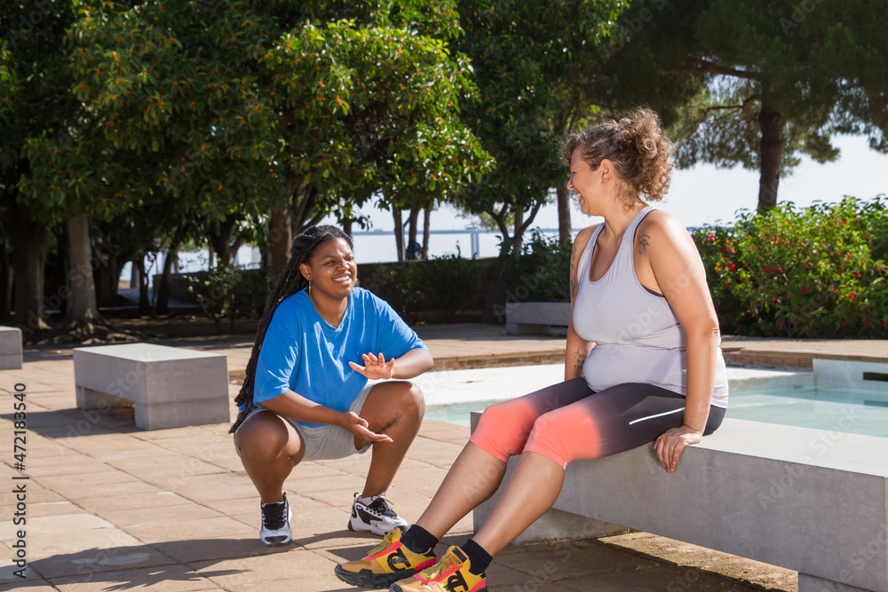 Obraz premium Friendly plus size women resting after training. Chubby women of different nationalities in sport clothes sitting on bench, talking. Sport, body positive concept