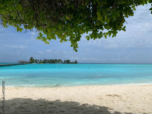 A view from under the branches of a tree to the shores of the Indian Ocean with white sand, azure water and the neighboring island, to which a wooden platform leads in the Maldives © Elena