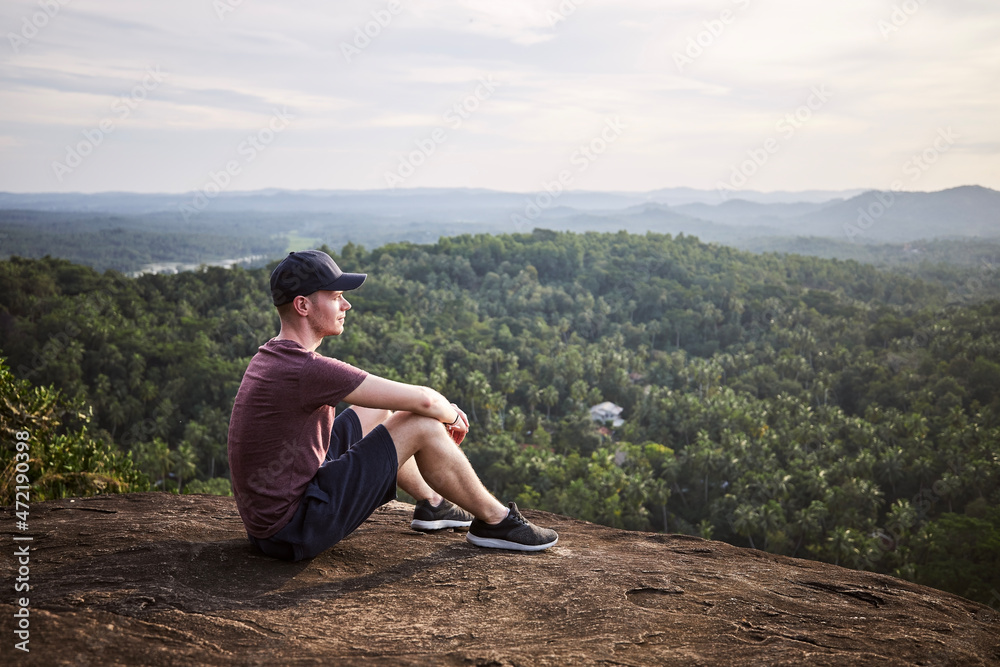 Pensive man resting at the edge of a rock and looking at view. Landscape with tropical rainforest in Sri Lanka..