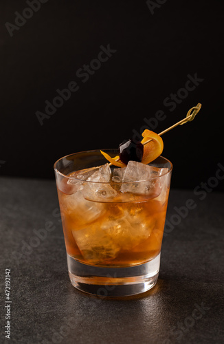 Coffee Old Fashioned Cocktail with Cherry and Orange. Cold bright cocktail on a black background. Lumps of ice in a glass. Party aesthetics