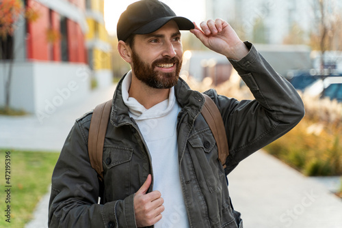 Portrait view of the bearded man wearing cap smiling toothy while standing at the street with backpack in front of the camera