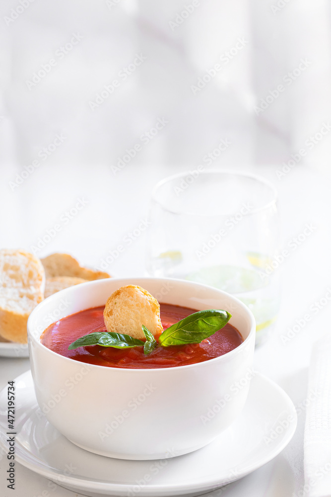 Tomato soup in white bowl decorated with bread crumbs, fresh basil and herbs on the restaurant table with water glass and portion of bread . Tomato soup in white bowl decorated with bread crumbs, fresh basil and herbs on the restaurant table with water glass and portion of bread .