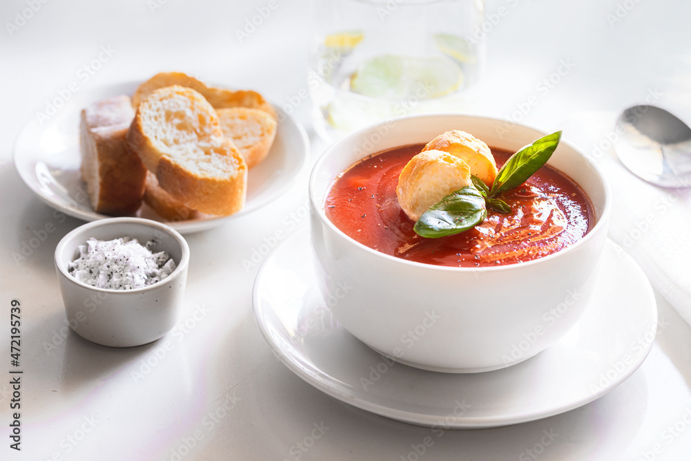 Tomato soup in bowl decorated with bread crumbs, fresh basil and herbs on the restaurant table with water glass and portion of bread . Tomato soup in bowl decorated with bread crumbs, fresh basil and herbs on the restaurant table with water glass and portion of bread .