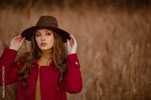 Outdoor autumn portrait of elegant woman wearing stylish brown hat, red marsala color classic coat posing in nature. Model looking aside. Copy, empty space for text