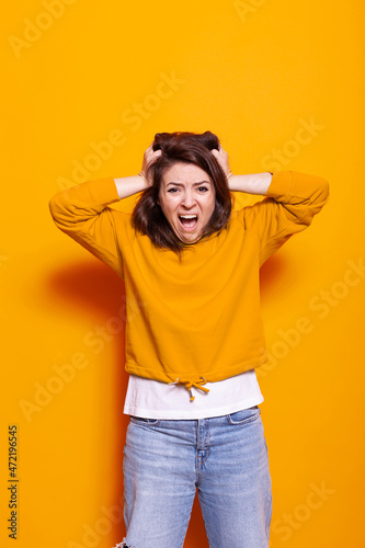 Desperate woman screaming and pulling hair with hands in studio. Young person with displeased and dramatic expression on face, feeling irritated while standing over orange background © DC Studio