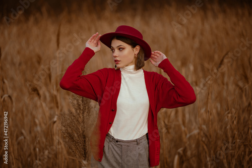 Elegant woman wearing stylish red marsala color hat, white turtleneck, orange cardigan posing in nature. Model looking aside. Outdoor autumn portrait. Copy, empty space for text photo