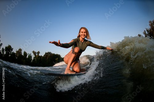 beautiful woman riding the wave while sitting on surf style wakeboard with arms outstretched to the sides
