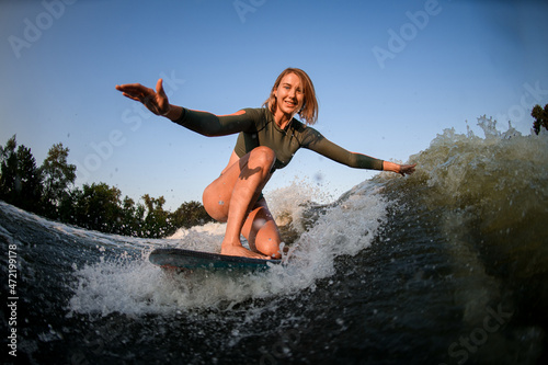 blonde woman riding the wave while sitting on surf style wakeboard with arms outstretched to the sides