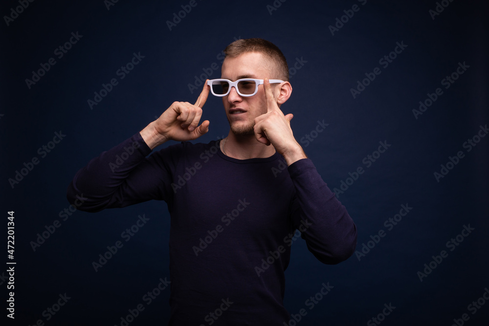 Smiling young man in a blue sweater and white glasses posing on a blue background, straightens his glasses and looks to the side Smiling young man in a blue sweater and white glasses posing on a blue background, straightens his glasses and looks to the side