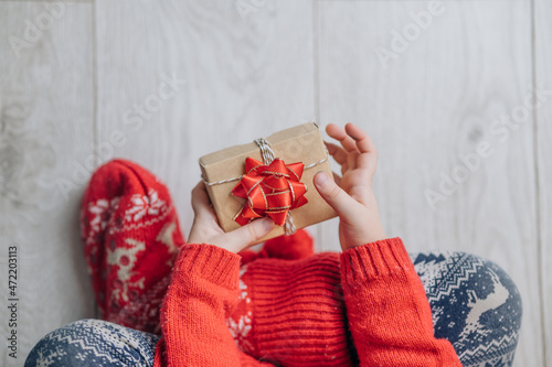 Child hands holding one gift box wrapped in kraft paper tied with red ribbon boow over wooden background. Top view, place for text. Holiday concept. photo