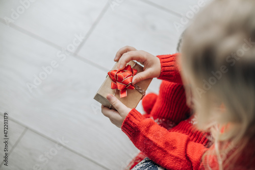 Child hands holding one gift box wrapped in kraft paper tied with red ribbon boow over wooden background. Top view, place for text. Holiday concept. photo