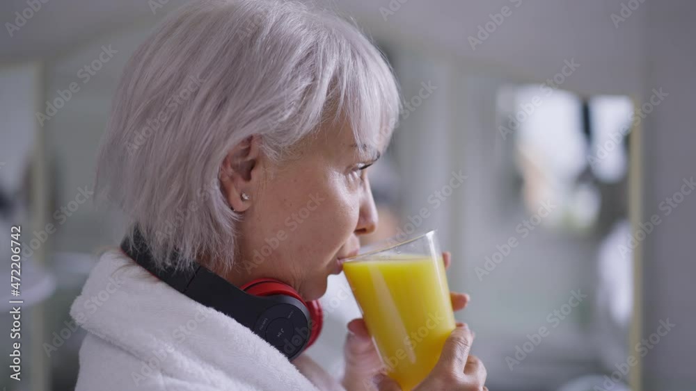 Side view portrait confident Caucasian mature woman drinking orange juice in the morning standing in bathroom at home. Grey-haired beautiful middle aged lady enjoying healthful drink indoors Side view portrait confident Caucasian mature woman drinking orange juice in the morning standing in bathroom at home. Grey-haired beautiful middle aged lady enjoying healthful drink indoors