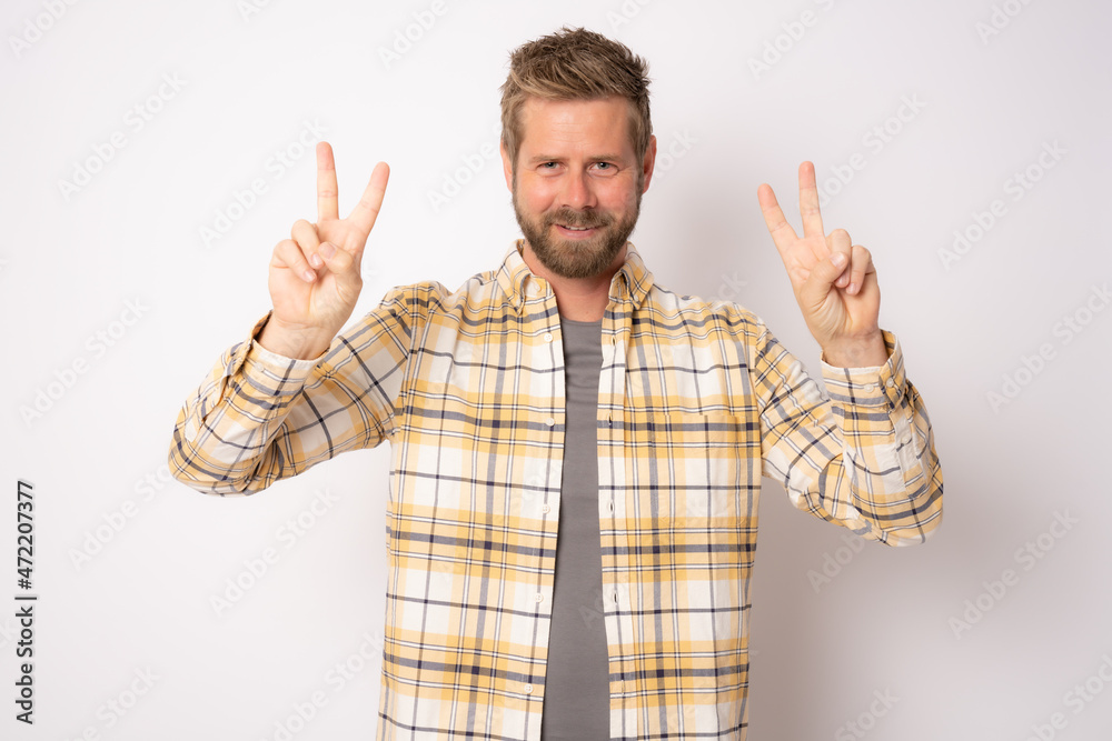 Handsome young man with beard wearing plaid shirt smiling looking to the camera showing fingers doing victory sign. Handsome young man with beard wearing plaid shirt smiling looking to the camera showing fingers doing victory sign.