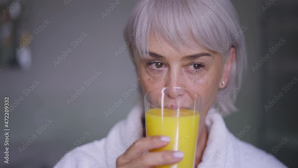 Front view portrait confident middle aged woman drinking orange juice in slow motion looking away. Slim beautiful Caucasian mature lady standing at home in the morning enjoying taste of healthy drink Front view portrait confident middle aged woman drinking orange juice in slow motion looking away. Slim beautiful Caucasian mature lady standing at home in the morning enjoying taste of healthy drink