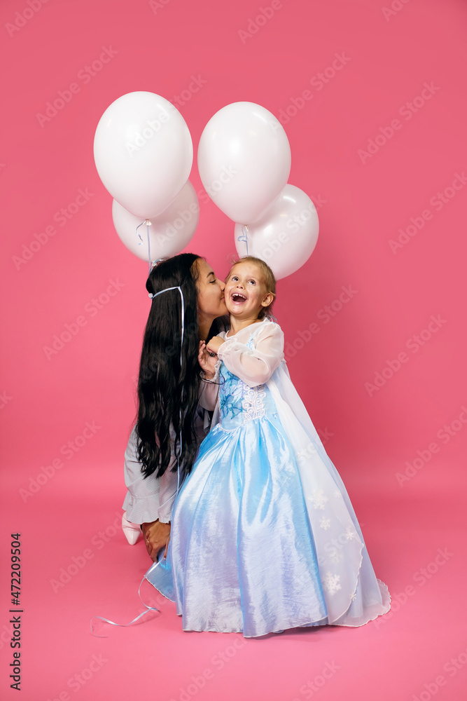 Young happy brunette mother kissing her charming smiling birthday girl daughter with white balloons attached to her hair. Attractive mother and her lovely daughter in blue dresses on pink background Young happy brunette mother kissing her charming smiling birthday girl daughter with white balloons attached to her hair. Attractive mother and her lovely daughter in blue dresses on pink background
