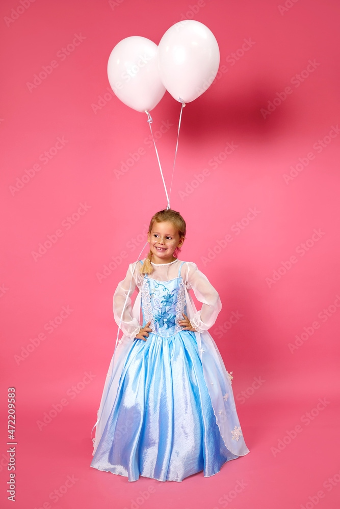 Beautiful happy little girl in a blue dress posing on a pink background with white balloons. Festive mood and birthday photo session. White balloons are attached to the hair of a little girl Beautiful happy little girl in a blue dress posing on a pink background with white balloons. Festive mood and birthday photo session. White balloons are attached to the hair of a little girl