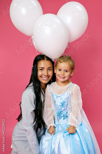 Young happy mother and her charming smiling daughter birthday girl with white balloons on a pink background. Attractive mother and her lovely daughter in blue dresses on pink background