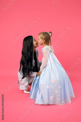 Young beautiful mother and her daughter are dressed in blue dresses and kiss on a pink background. Mom and daughter are posing for the photographer on a pink background