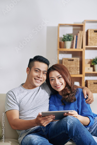 Portrait of positive young Asian couple using digital tablet and embracing together in living room