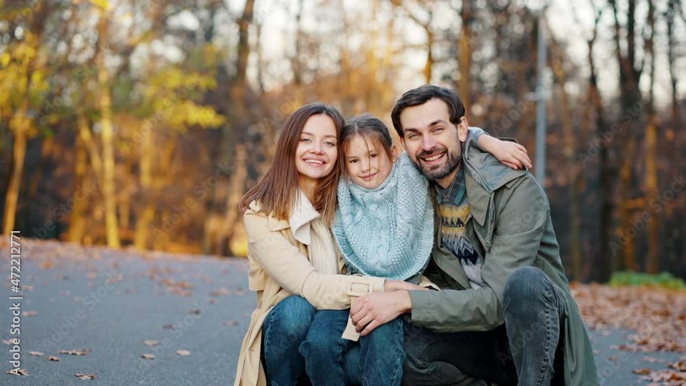 Young smiling mom, dad and little daughter are hugging while sitting squatting in autumn park. Happy family. Close up, slow motion Young smiling mom, dad and little daughter are hugging while sitting squatting in autumn park. Happy family. Close up, slow motion