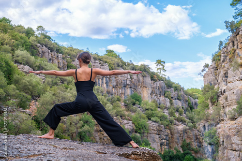 Back view full body of anonymous barefoot slender female practicing Virabhadrasana posture while standing on rocky cliff during yoga training in mountainous terrain