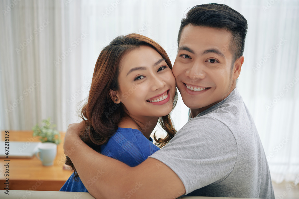 Portrait of cheerful young Asian couple looking back and embracing on sofa in living room Portrait of cheerful young Asian couple looking back and embracing on sofa in living room