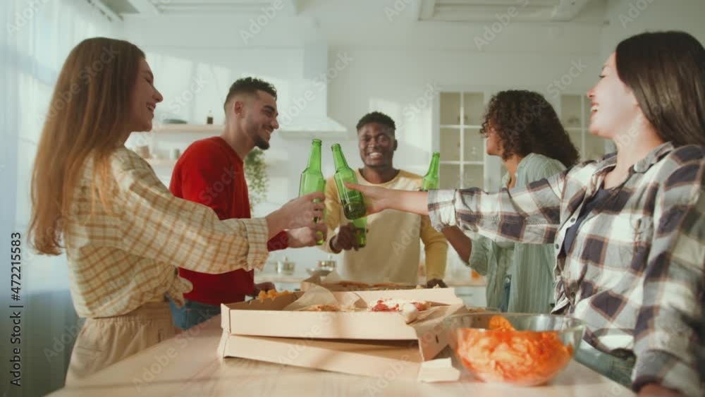 Group of happy diverse young friends clinking with beer bottles over dinner table, enjoying weekends together, zoom out Group of happy diverse young friends clinking with beer bottles over dinner table, enjoying weekends together, zoom out
