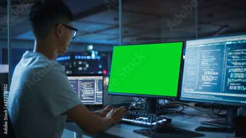 Night Office: Young Japanese Man in Working on Green Screen Chroma Key Desktop Computer. Team of Programmers Typing Code, Creating Modern Software, e-Commerce App Design, e-Business Programming