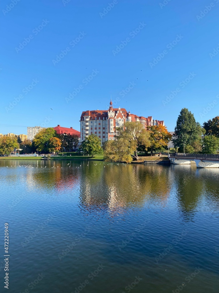 Fototapeta premium Breathtaking view to The Lower Pond and a park near it in Kaliningrad city in Russia in the golden autumn season. Colorful trees and clouds reflect in the lake waters