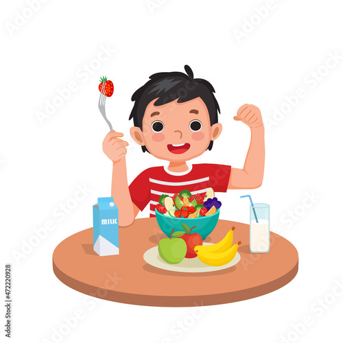 Cute little boy eating healthy fruits and vegetables showing fork with strawberry and his strong muscle arm fist.