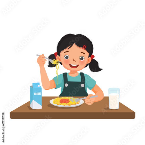 Cute little girl feeling excited eating delicious spaghetti using fork and spoon with a glass of milk