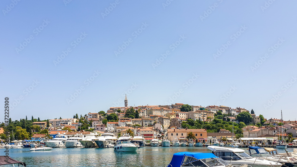 A view on the harbour in Vrsar. There are a lot of boats anchored to the shore. In the back, there is the city center, with a tall church tower. Clear and sunny day. Busy day in the harbour.