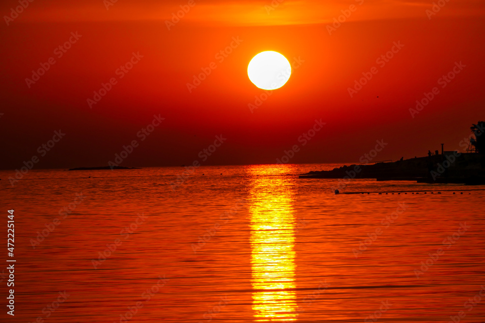 Romantic sunset by a beach. The sun sets over the horizon. The sun beams reflecting in the calm sea waters. There is an island on the side. Few birds flying around. The sky turns yellow and orange